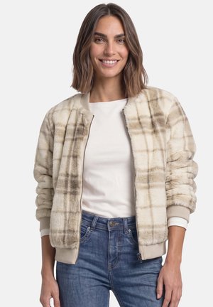 Kvinde smilende iført en beige ternet bomberjakke i imiteret pels over en hvid skjorte og blå jeans med hænderne afslappet ved siden.