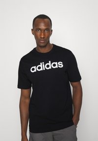 Melna kokvilnas T-krekls ar īsām piedurknēm un apaļu kakla izgriezumu, uz kuras ir liels balts "adidas" logo krūtīs. Gluda tekstūra.