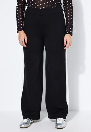 Henkilö, jolla on mustat leveälahkeiset housut, hopeiset lenkkarit ja musta pitkähihaisen paita, jossa on vaaleanpunaisia sydänkuvioita, seisoo valkoista seinää vasten.