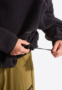 Μαύρο πουλόβερ από φλίς με υφή, με τσέπες με φερμουάρ και ρυθμιστικό με κορδόνι στο τελείωμα. Φορεμένο πάνω από ελιά σορτς.