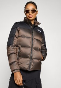 Donzen pufferjack met een kleurblokontwerp in brons en zwart, voorzien van ritssluitingen en een hoge kraag. Logo op de borst.