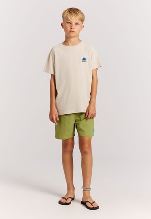 Beige katoenen T-shirt met een kleine blauwe en groene zonafbeelding, gecombineerd met groene shorts en zwarte teenslippers. Eenvoudig, casual ontwerp.