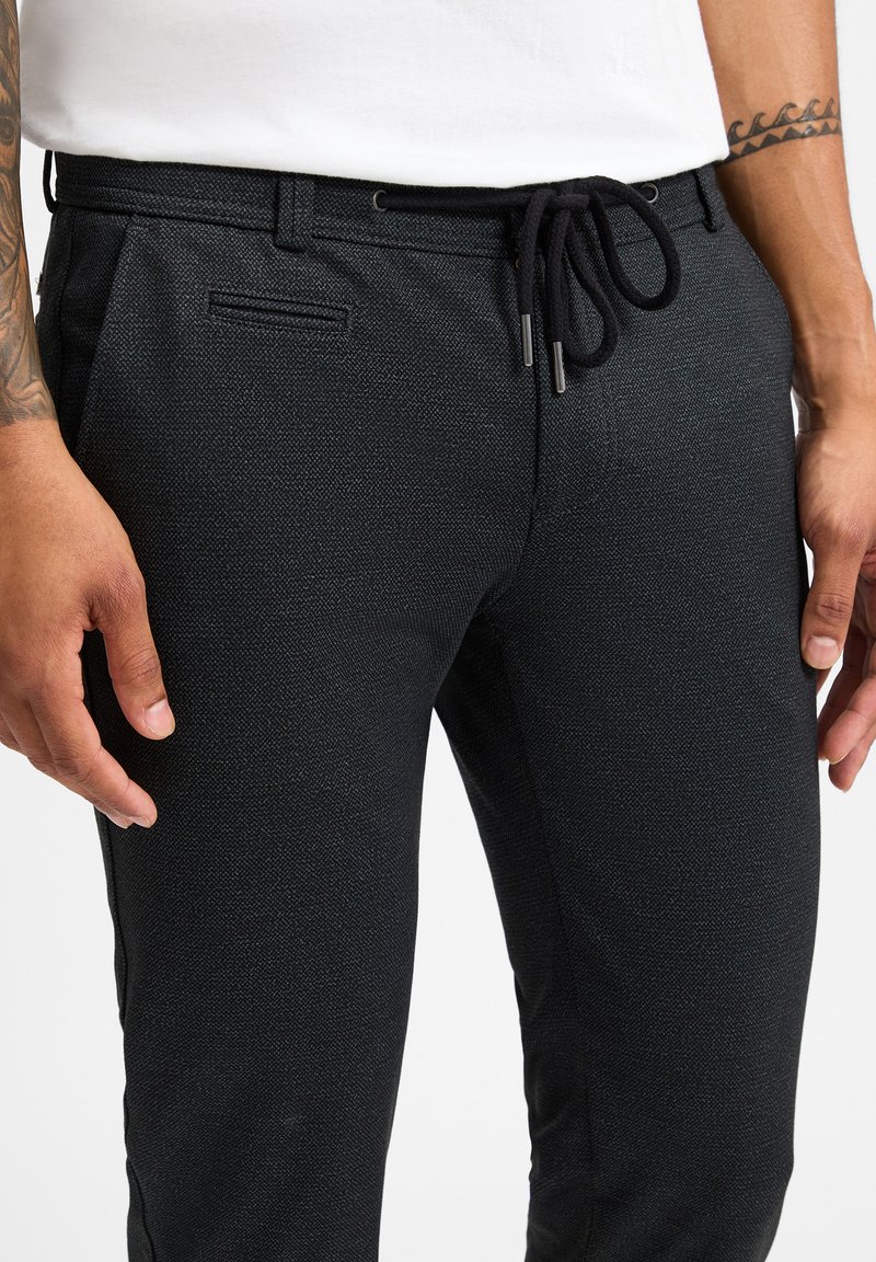 Schwarze Hose aus strukturiertem Stoff, mit weitem Bund und Seitentasche, in taillierter Silhouette und subtilen Muster.