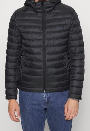 Mand iført en sort polstret jakke med lynlås og hætte samt blå jeans, stående med armene afslappet ned langs siden.