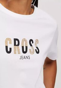 Biała bawełniana koszulka z napisem "CROSS" w czerni z złotymi ćwiekami oraz "JEANS" poniżej w czerni. Okrągły dekolt, krótkie rękawy.
