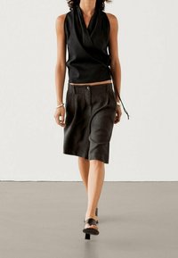 Schwarzes ärmelloses Wickeltop, kombiniert mit dunklen, knielangen Shorts. Das Modell trägt schwarze Sandalen mit offenen Zehen und präsentiert ein minimalistisches Design.