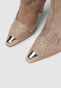 Botas de tobillo de ante marrón con punta afilada, que presentan acentos metálicos en la punta y un intrincado detalle de cuentas a lo largo del borde superior.