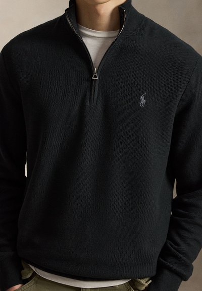 Homme portant un pull noir à fermeture éclair quatre-quarts avec tissu texturé et petit logo brodé sur la poitrine gauche, superposé à une chemise blanche.
