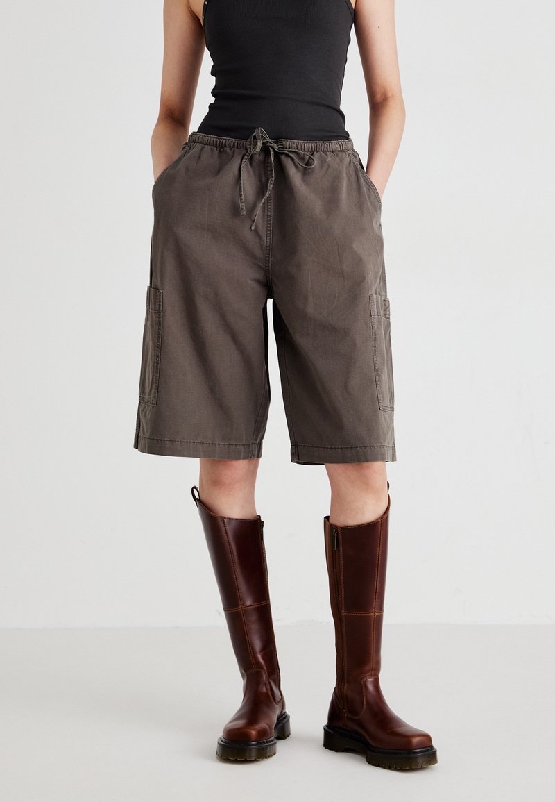 Personne portant un short marron ample jusqu'aux genoux avec des poches latérales, un haut noir sans manches et de grandes bottes en cuir marron. Mains dans les poches.