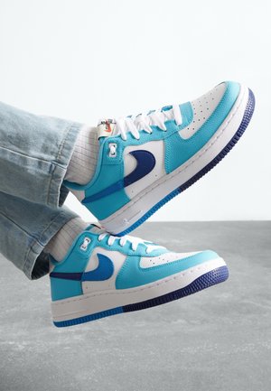 Person iført hvide sokker og blå-hvide Nike-sneakers med mørkeblå Swoosh, lyseblå accenter og lyseblå jeans.