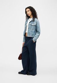 Γυναίκα με σγουρά μαλλιά φοράει ελαφρύ τζιν μπουφάν, λευκό crop top, φαρδιά σκούρα μπλε παντελόνια, μαύρα παπούτσια και κρατάει μια μικρή καφέ τσάντα.