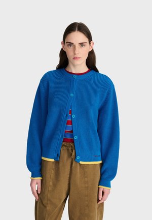 Jeune femme portant un cardigan bleu clair boutonné avec une bordure jaune par-dessus une chemise rayée rouge et bleue et un pantalon marron, debout devant un fond blanc.