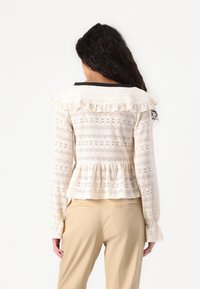 Krajkový peplum top v krémovej farbe s volánovým golierom a dlhými rukávmi. Obsahuje zložitý kvetinový vzor a čierny lem na výstrihu.