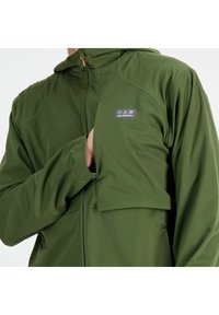 Grüne Zip-Jacke aus glattem, leichtem Stoff. Mit einem Logo-Patch auf der Brust und einer Kapuze mit durchgehendem Reißverschluss.