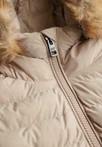 Beige gewatteerde jas met een doorgestikt ontwerp, voorzien van een zilveren rits, "Superdry" merknaam en een accent van faux bont aan de capuchon.