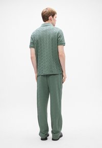 Ung man som står vänd bort, klädd i en matchande grön texturerad pikétröja och vida byxor med svarta skor mot en vit bakgrund.