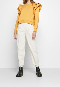 Žlutá mikina s volánovými rukávy, zkombinovaná s krémovými plisovanými kalhotami a černými kotníkovými botami. Hladká textura a pohodlný střih.