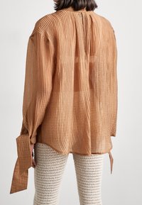 Camicia lunga a maniche con texture marrone, con pieghe e lacci laterali, caratterizzata da uno scollo a chiave sul retro. Abbinata a pantaloni bianchi all'uncinetto.