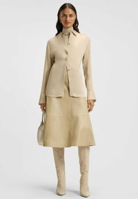 Beige Langarmhemd mit Kragen und Knöpfen, kombiniert mit einem passenden A-Linien-Rock. Das Model trägt hohe beige Stiefel und hält eine kleine Tasche.