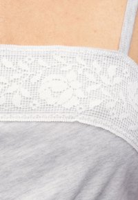 Κοντινή λήψη ενδύματος από ανοιχτό γκρι ύφασμα με λευκή δαντέλα με λουλούδια και λεπτό ιμάντα ώμου πάνω σε γυμνό δέρμα.