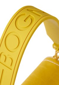 Geel rubberen handvat van een handtas met reliëflettering en een gladde textuur, bevestigd aan een leren lichaam. Heldere kleur, modern ontwerp.