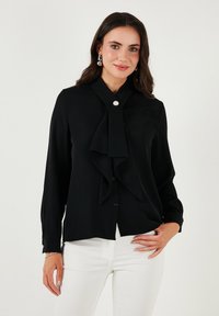 Black blouse with a high collar and draped tie detail, featuring button closure and long sleeves. Smooth fabric, paired with white trousers.