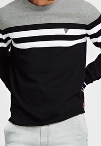 Sweatshirt rayé noir et gris avec un col rond, des rayures horizontales blanches, et un petit logo triangulaire sur le côté gauche de la poitrine. Texture douce.