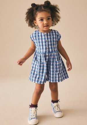 Niebiesko-biała sukienka w kratę bez rękawów z guzikami z przodu, dwoma bocznymi kieszeniami i kwiatowymi akcentami. Noszona z niebieskimi wysokimi trampkami.