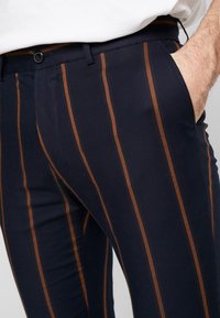 Navy blue trousers with brown vertical stripes, made from fabric with a smooth texture. Features a button closure and side pocket.