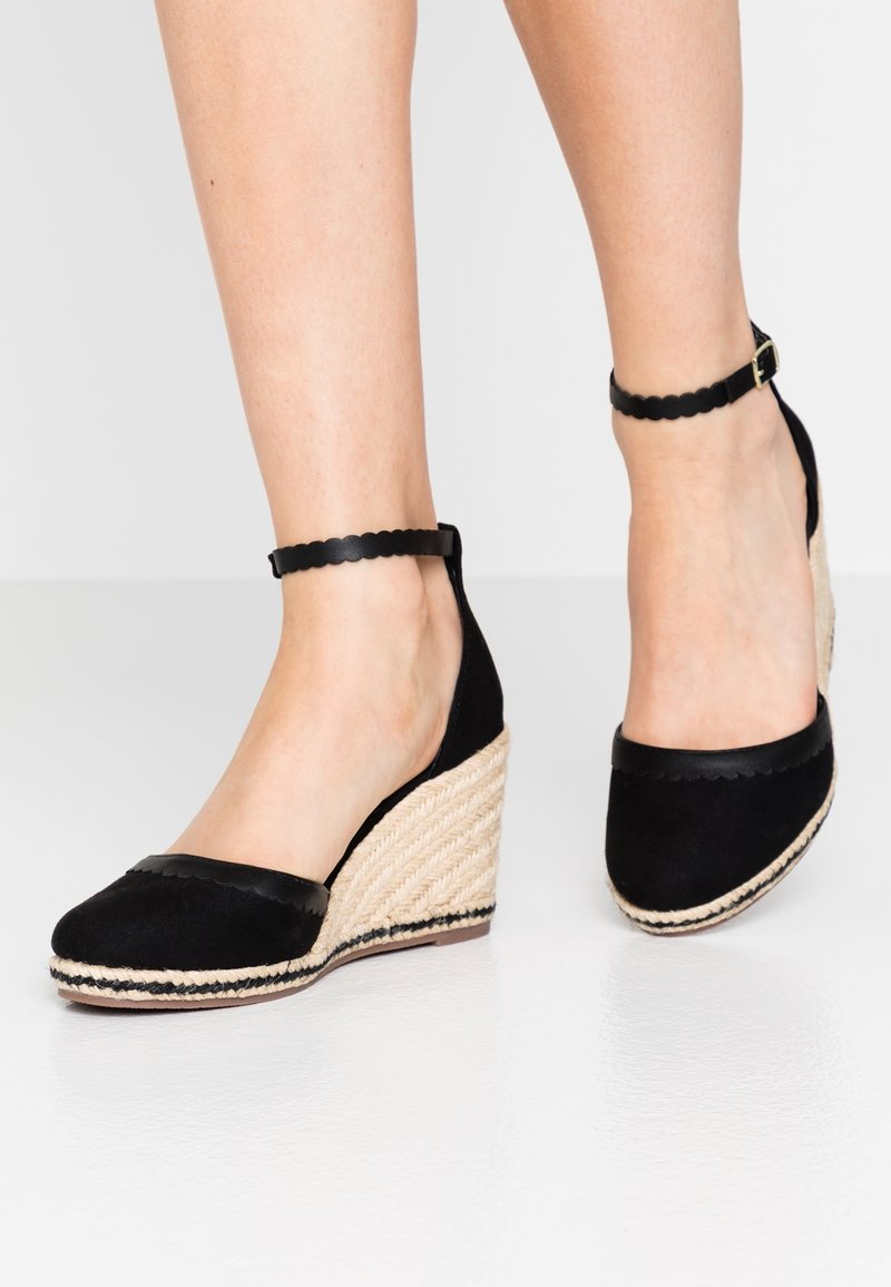 Espadrilles compensées noires avec brides autour de la cheville, bords festonnés et talons en corde tressée portées pieds nus.