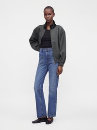 Femme portant une veste zippée noire, un top noir, un jean bleu taille haute avec des poches plaquées et des chaussures plates noires, debout sur fond blanc.