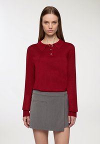 Czerwona, długa koszulka polo z kołnierzykiem i trzema brązowymi guzikami, w połączeniu z szarym, strukturalnym mini spódnicą z kieszonką boczną.