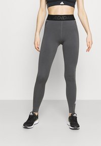 Šedé sportovní legíny s širokým černým pasem, na kterém je logo "ADIDAS", hladký povrch a přiléhavý design pro pohodlí a flexibilitu.