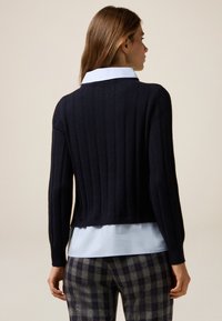 Marineblauer gerippter Strickpullover mit einem verkürzten Design, der lange Ärmel und einen runden Ausschnitt hat, über einem hellblauen Hemd mit Kragen getragen.