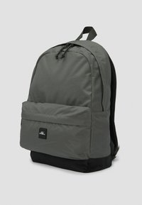 Mochila de tela gris con parte superior redondeada, bolsillo frontal y sección inferior negra. Correas ajustables y detalle de cierre con cordón.