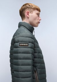 Gewatteerde jas in donkergroen, met een hoge kraag en ritssluiting. Bevat een merkpatch op de linkermouw en textuur met horizontale lijnen.
