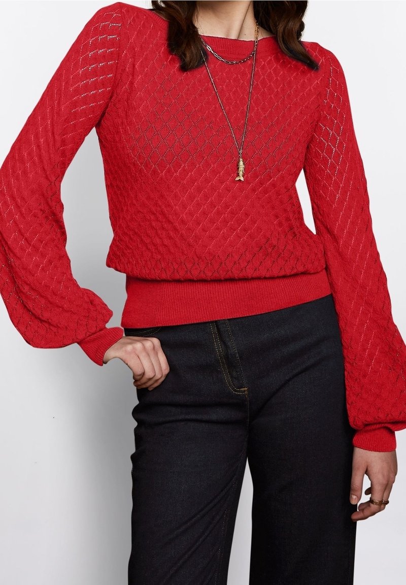 Czerwony sweter dziany w teksturowany wzór diamentowy, z prążkowanym dołem i bufiastymi rękawami, zestawiony z ciemnymi denimowymi spodniami.