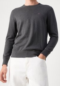 Homme portant un pull à manches longues gris foncé et un pantalon blanc, une main dans la poche, debout devant un fond clair uni.