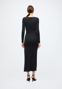 Zwarte jurk met lange mouwen, aansluitend silhouet, gladde stof, tot op de knie, sluiting aan de achterkant met een druppelvormige opening, gecombineerd met high-heeled sandalen met bandjes.