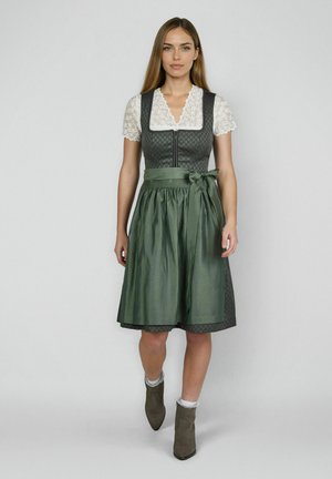 Vrouw in een groene dirndl-jurk met een witte kanten blouse en een bijpassende groene schort, lopend tegen een effen grijze achtergrond.