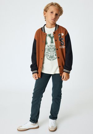 Brun og sort varsity jakke, hvid t-shirt med grøn grafik, mørke jeans og hvide sneakers. Jakken har kontrasterende ærmer og stribede manchetter.