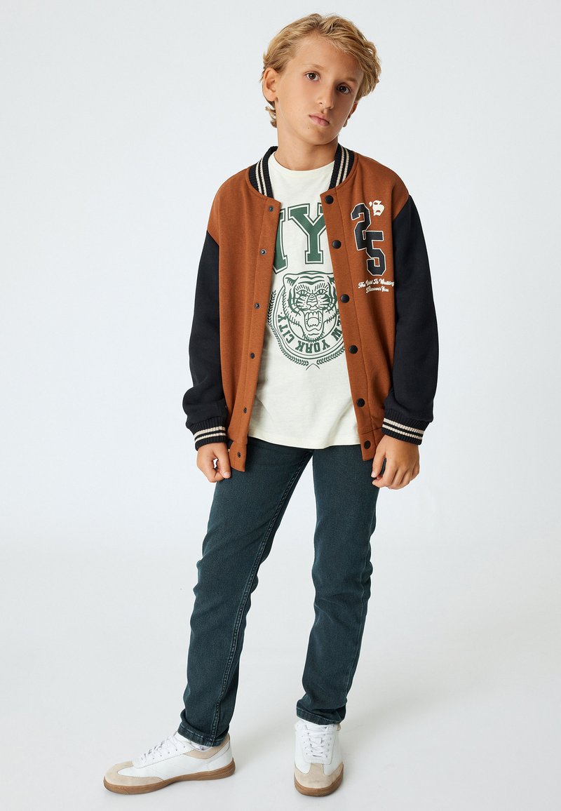 Brun og sort varsity jakke, hvid t-shirt med grøn grafik, mørke jeans og hvide sneakers. Jakken har kontrasterende ærmer og stribede manchetter.