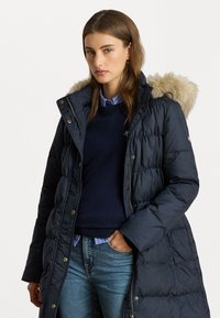 Hosszú sötétkék pufi kabát, prémes kapucnival, tűzött mintával és aranyszínű részletekkel. Sötét pulóverrel és kék csíkos inggel párosítva.