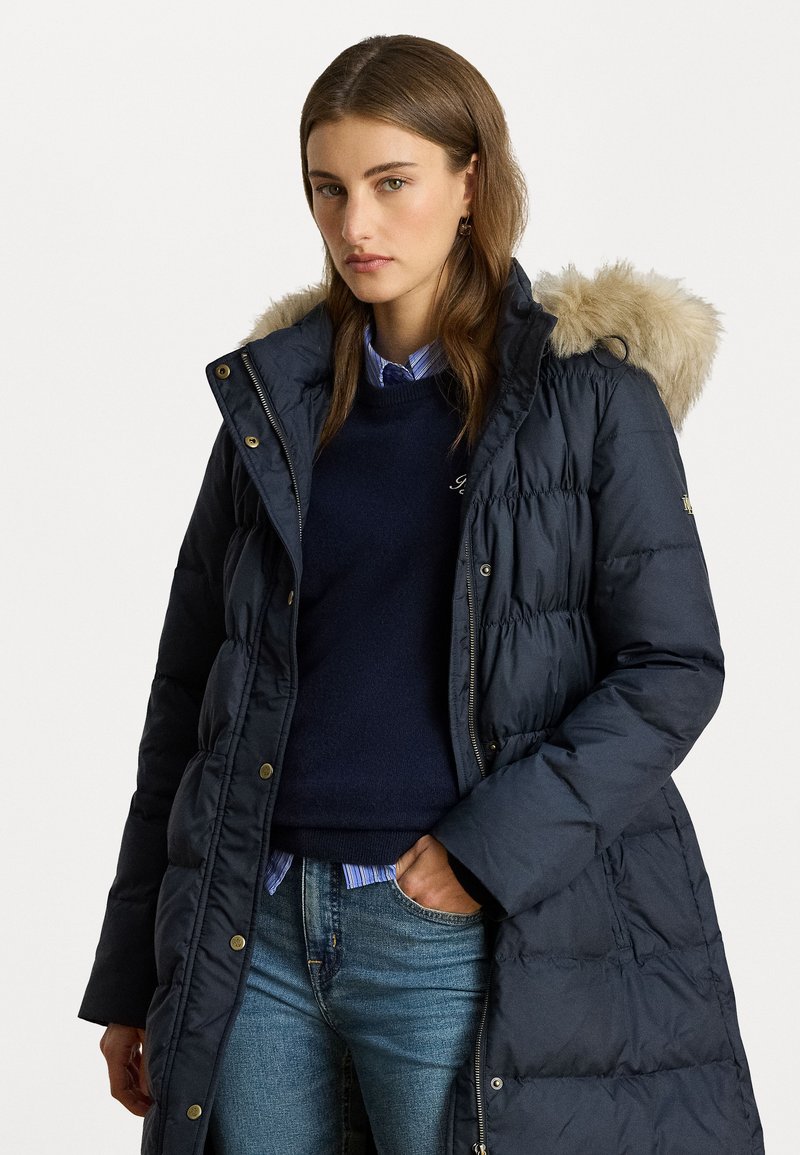 Hosszú sötétkék pufi kabát, prémes kapucnival, tűzött mintával és aranyszínű részletekkel. Sötét pulóverrel és kék csíkos inggel párosítva.