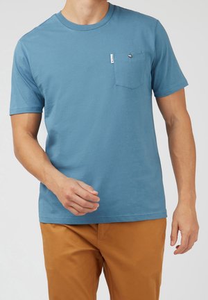 Mand iført en blå t-shirt med korte ærmer med en lille brystlomme og brune bukser foran en ensfarvet baggrund.