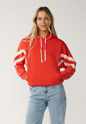 Vrouw met lang blond haar die een rode hoodie met witte strepen op de mouwen en lichtblauwe spijkerbroek draagt, staand met handen in de zakken.