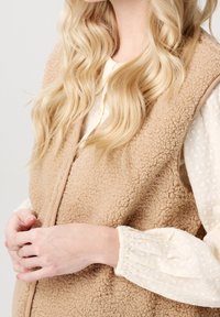 Beige ärmellose Weste aus Kunstfell mit strukturierter Oberfläche. Darüber getragen eine cremefarbene Langarmbluse mit gerafften Bündchen.