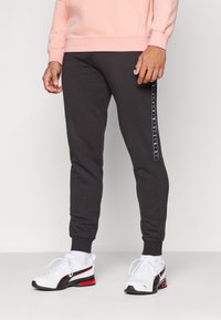 Donkergrijze sweatpants met een taps toelopende pasvorm, voorzien van een zijlogo-strijk. Gecombineerd met wit en zwarte sportschoenen. Gestructureerde stof.