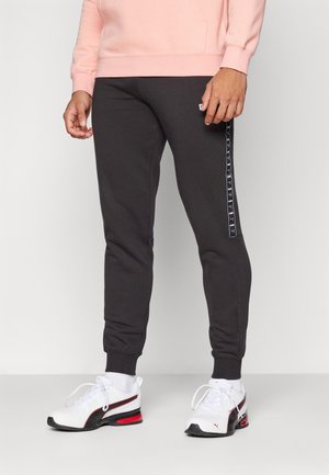 Dark grey sweatpants with a tapered fit, featuring a side logo stripe. Paired with white and black athletic shoes. Textured fabric.