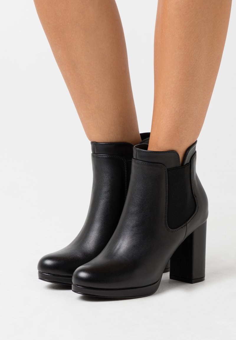 Bottes à cheville en cuir noir à talons carrés portées sur des jambes nues, sur fond blanc.